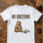 A white short-sleeve graphic T-shirt laid flat on a rustic wooden surface, featuring the text 'NO QUESTIONS just coffee' above an illustration of a capybara holding a mug with coffee beans and a broken branch nearby. A glass of iced coffee and scattered coffee beans appear as styled props around the shirt.