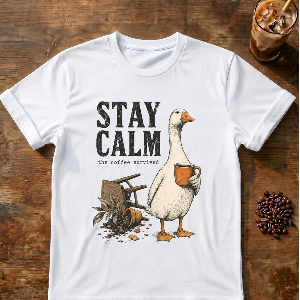 A white short-sleeve graphic T-shirt laid flat on a rustic wooden surface, featuring the words “STAY CALM” above an illustration of a goose holding an orange mug with coffee beans and a spilled coffee can nearby. A glass of iced coffee and a pile of coffee beans are placed around the shirt as styled props.