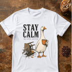 A white short-sleeve graphic T-shirt laid flat on a rustic wooden surface, featuring the words “STAY CALM” above an illustration of a goose holding an orange mug with coffee beans and a spilled coffee can nearby. A glass of iced coffee and a pile of coffee beans are placed around the shirt as styled props.
