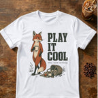 A white short-sleeve graphic T-shirt laid flat on a rustic wooden surface, featuring an illustration of a fox holding a mug next to the text 'PLAY IT COOL' and smaller text 'they know nothing.' Coffee-themed props, including an iced coffee glass and coffee beans, are placed around the shirt.