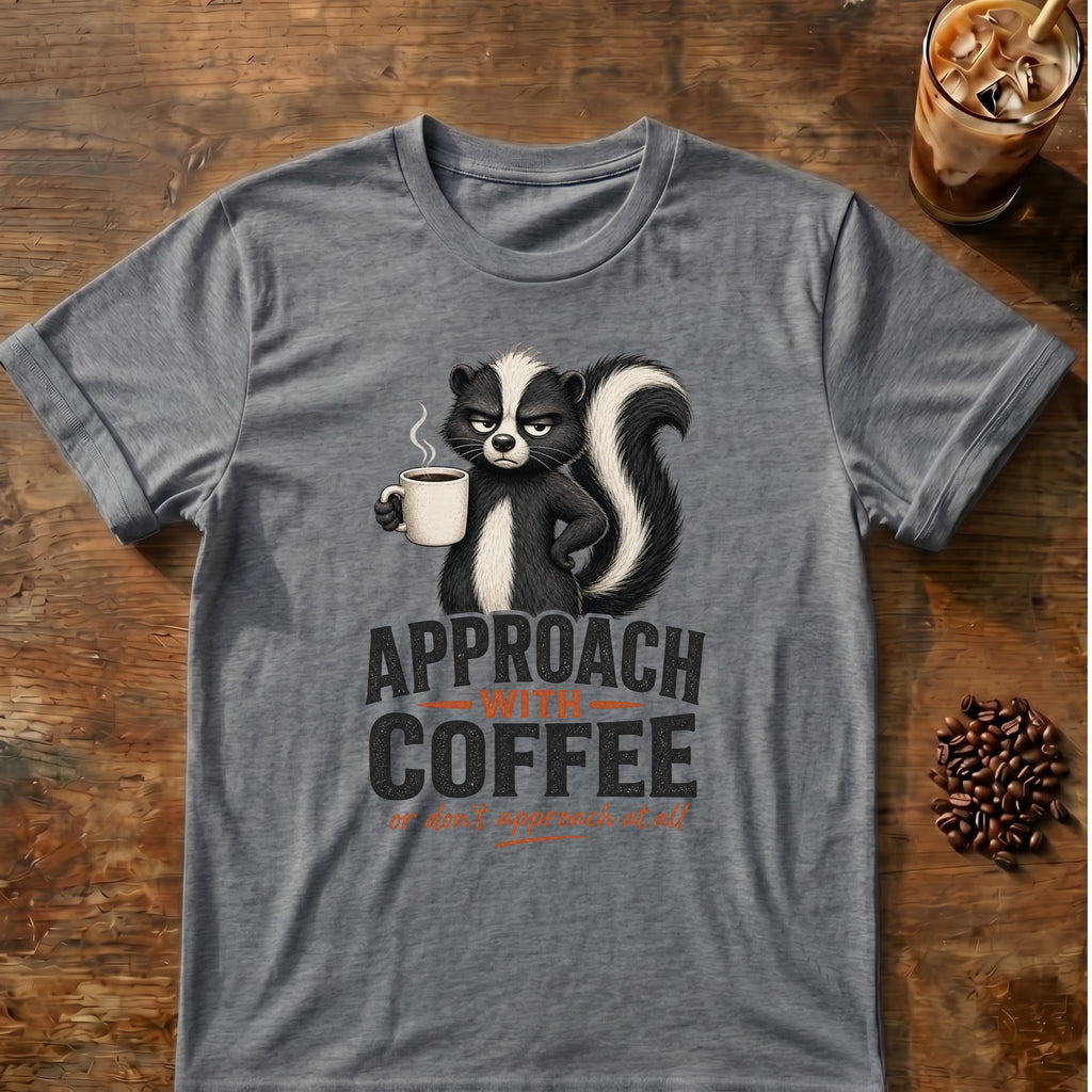 Grumpy skunk holding a steaming coffee mug above bold text reading Approach With Coffee, with orange script below saying or don’t approach at all. Graphite Heather