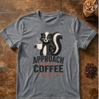 Grumpy skunk holding a steaming coffee mug above bold text reading Approach With Coffee, with orange script below saying or don’t approach at all. Graphite Heather