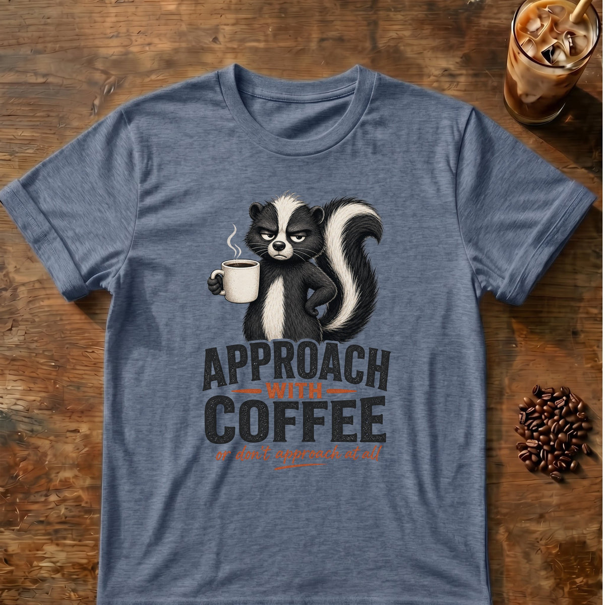 Grumpy skunk holding a steaming coffee mug above bold text reading Approach With Coffee, with orange script below saying or don’t approach at all. Heather Indigo