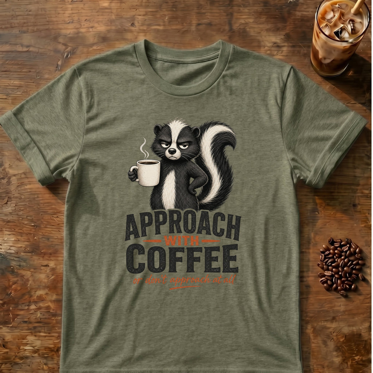 Grumpy skunk holding a steaming coffee mug above bold text reading Approach With Coffee, with orange script below saying or don’t approach at all. Heather Military Green