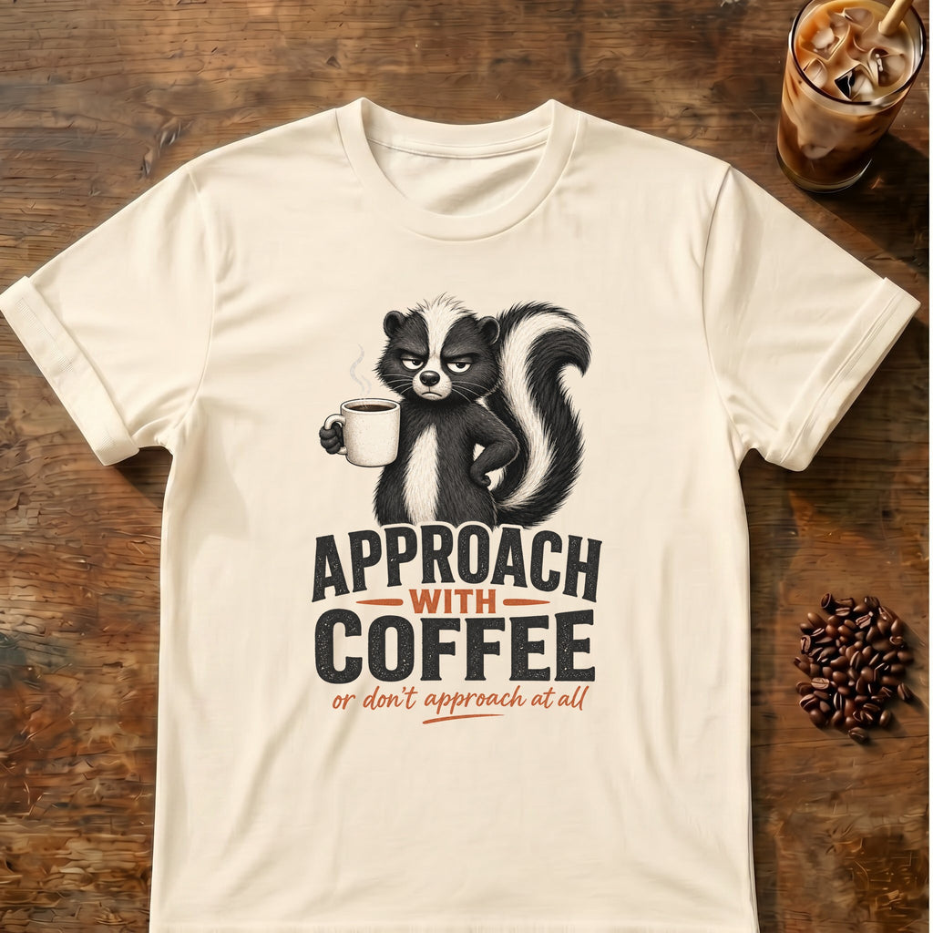 Grumpy skunk holding a steaming coffee mug above bold text reading Approach With Coffee, with orange script below saying or don’t approach at all. Natural