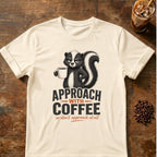 Grumpy skunk holding a steaming coffee mug above bold text reading Approach With Coffee, with orange script below saying or don’t approach at all. Natural